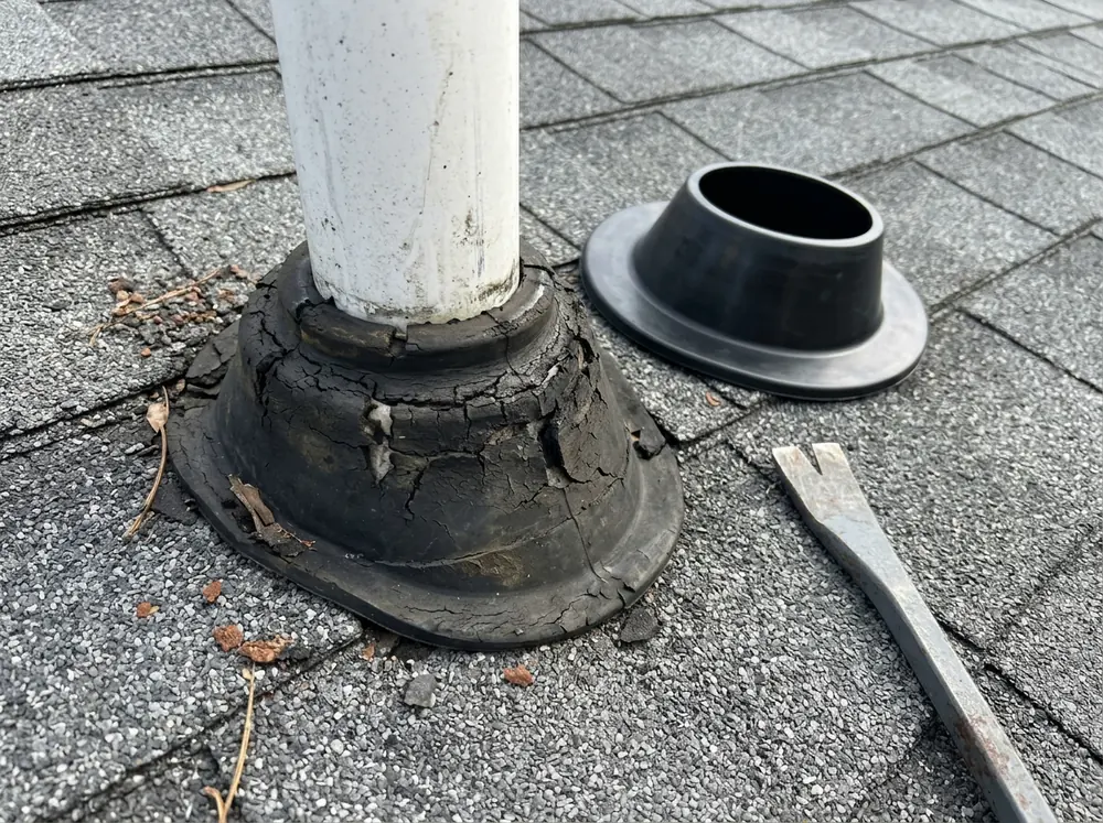Close view of a cracked rubber vent boot around a plumbing pipe on an asphalt shingle roof, with a new boot beside it