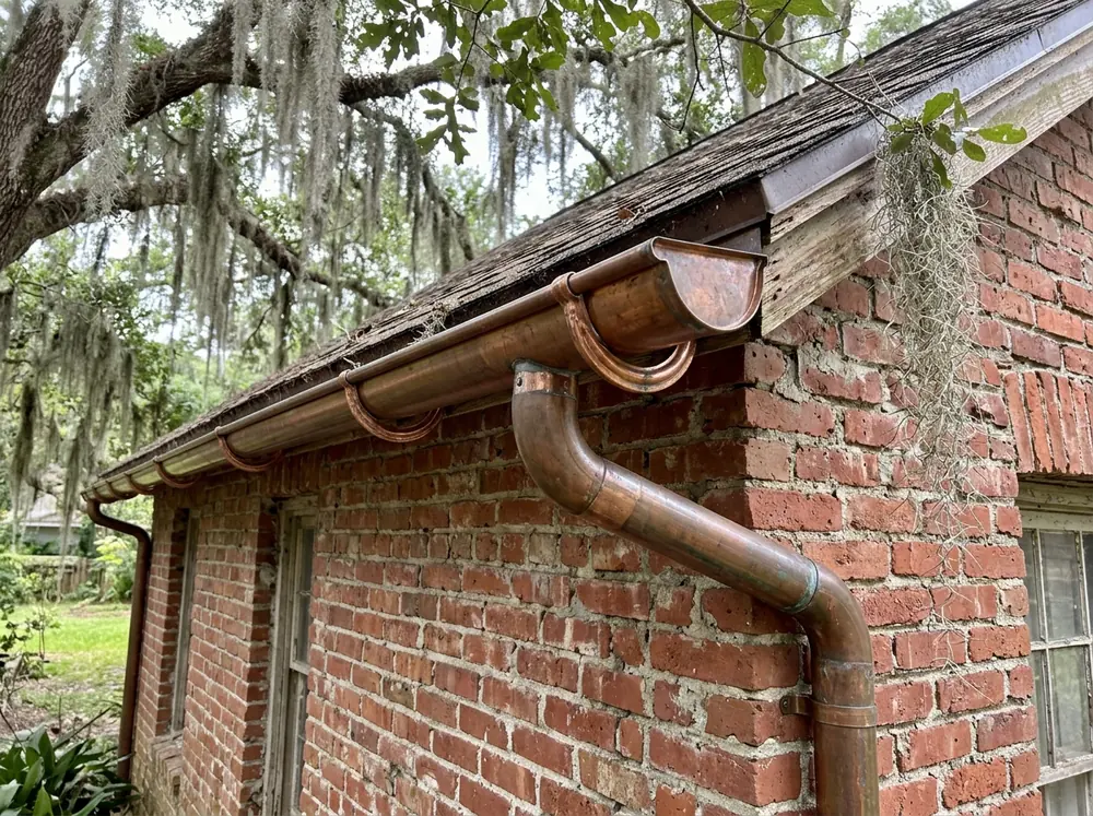 Half-round gutter with round downspout along the eave of an older red brick house shaded by a live oak