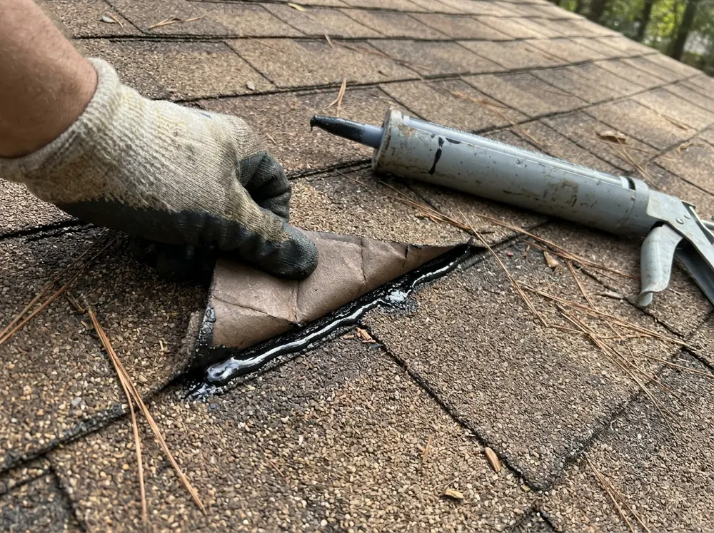Roofer's gloved hand pressing a lifted asphalt shingle tab back down over a bead of roofing sealant